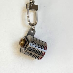 May include: A silver-toned necklace featuring a pendant resembling a combination lock. The lock has rotating dials with white numbers and red accents. The necklace includes a small clasp and a delicate chain.