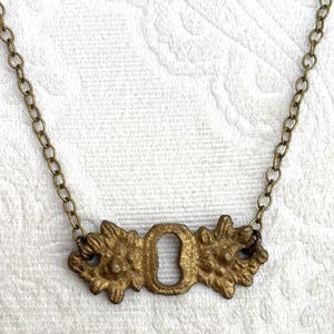 May include: A vintage-style brass necklace featuring a keyhole pendant. The pendant is intricately detailed with floral accents on either side of the keyhole. The chain is a matching brass color, creating a cohesive and antique aesthetic.