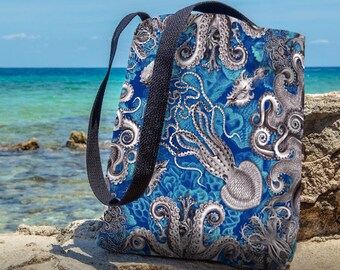May include: A blue tote bag with a white and gray octopus design. The bag has black straps and is sitting on a rocky surface near the ocean.