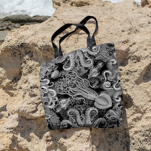 May include: A black tote bag with white octopus and sea life illustrations. The bag has black straps and is sitting on a rocky surface.