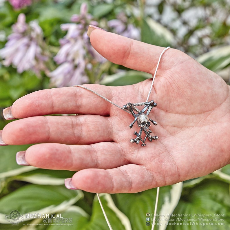 K&ouml;nnte beinhalten: Eine silberne Halskette mit einem Totenkopf- und Knochenkreuz-Anh&auml;nger. Der Anh&auml;nger ist detailliert und kunstvoll gearbeitet, mit einem realistischen Totenkopf und gekreuzten Knochen. Die Halskette ist an einer silbernen Kette.