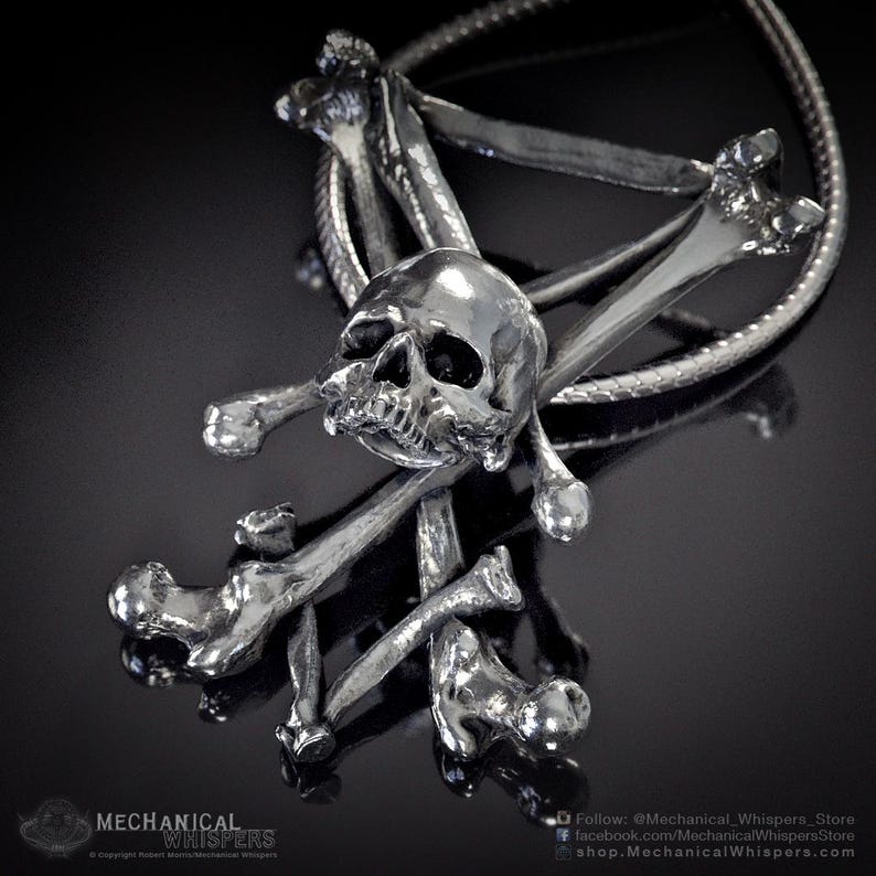K&ouml;nnte beinhalten: Eine silberne Halskette mit einem Anh&auml;nger in Form eines Totenkopfs mit gekreuzten Knochen. Der Totenkopf ist detailliert mit einem markanten Kiefer und Augenh&ouml;hlen. Die gekreuzten Knochen bestehen aus mehreren Knochensegmenten.