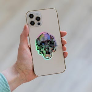 以下が含まれることがあります： 背面にホログラフィックなスカルステッカーが貼られた白いiPhone。ステッカーは虹色で、緑、青、紫の色合いです。