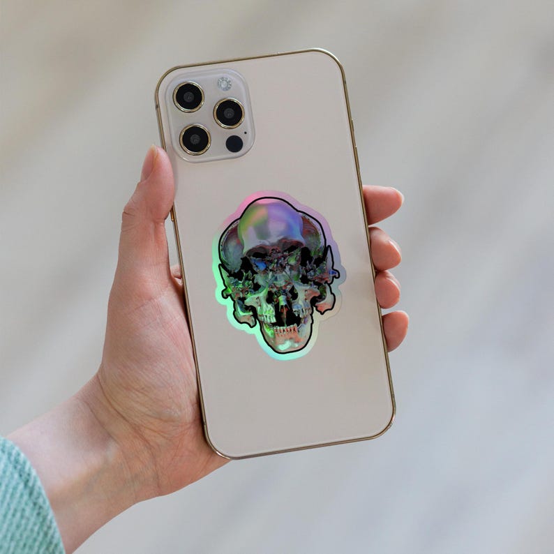 以下が含まれることがあります： 背面にホログラフィックなスカルステッカーが貼られた白いiPhone。ステッカーは虹色で、緑、青、紫の色合いです。