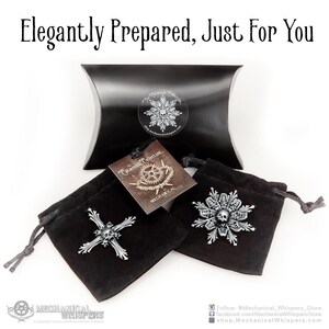 K&ouml;nnte beinhalten: Zwei schwarze Samtbeutel mit silbernen Totenkopf- und Schneeflockenmotiven aus Metall. Die Beutel sind in einer schwarzen Kissenbox mit dem Text "Catacomb Collection" und "Mechanical Whispers" verpackt.