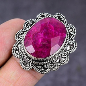 May include: A ring featuring a large, oval-shaped, faceted magenta gemstone set in a decorative sterling silver frame. The metalwork displays intricate scroll patterns and beaded edges surrounding the central stone, which has the text Silverstores.
