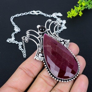 Peut inclure: Un collier avec un pendentif en argent orné d'une grande pierre rouge facetée en forme de larme. Le pendentif est entouré de filigranes argentés complexes et d'une petite pierre noire.