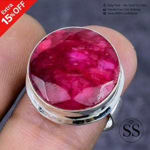 May include: A large, round ruby gemstone ring with a silver band. The ruby is a deep, vibrant red color and has a faceted surface. The ring is held by a person's finger. The image also includes the text "Extra 15% OFF" and "Silverstores".