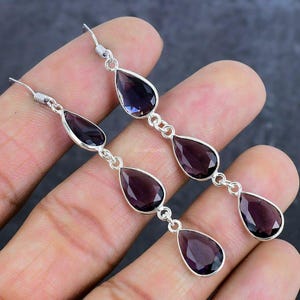 May include: A pair of sterling silver dangle earrings with three teardrop-shaped amethyst gemstones. The earrings are approximately 2 inches long.