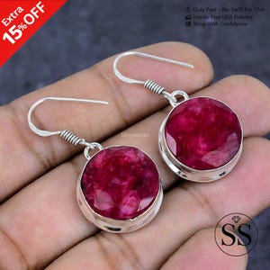 May include: Silver earrings with large, round, faceted, deep red gemstones. The earrings have a simple, elegant design with a silver hook and a decorative coil detail. The image also shows the text "Extra 15% OFF".