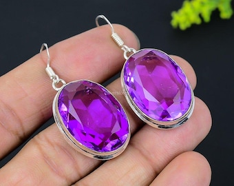 May include: A pair of silver-toned drop earrings featuring large, oval-cut, vibrant purple gemstones. The gemstones are set in a bezel setting, and the earrings have a simple hook closure. The earrings are displayed against a dark background.