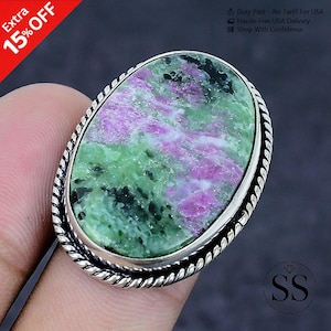 May include: An oval ring featuring a large, polished ruby zoisite gemstone with vibrant green and magenta-pink inclusions. The stone is set in a silver metal bezel with a decorative twisted rope-style border around the outer edge of the setting.