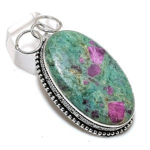 Puede incluir: Un colgante de plata con una piedra ovalada verde y rosa. La piedra preciosa tiene una superficie texturizada con inclusiones rosas y verdes. El colgante tiene una argolla de plata con un borde decorativo.