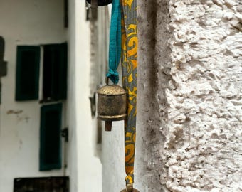 May include: Three gold bells hanging from a white wall. The bells are attached to colorful fabric ribbons with a floral pattern.