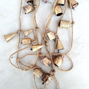 Pode incluir: Uma corda de sinos dourados pendurados em uma corda de juta natural. Os sinos são de vários formatos e tamanhos, alguns são redondos e outros têm forma de cone.