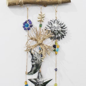 Op de afbeelding: Een wandhanger met hemelse motieven. Een houten staaf ondersteunt hangende ornamenten, waaronder een zon, maan en ster, samen met kralen en bellen. De zon en ster hebben lachende gezichten. Het kleurenschema omvat zilver, goud, blauw en groen.
