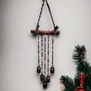 Op de afbeelding: Een kleurrijke kralenwindgong met een houten tak en metalen bellen. De windgong hangt aan een gevlochten koord met rode, witte en zwarte accenten.