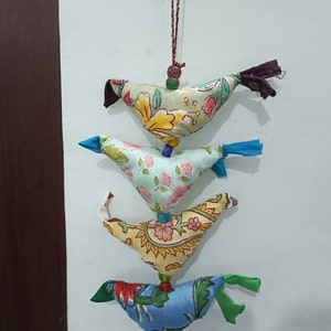 Könnte beinhalten: Eine Schnur mit vier bunten Stoffvögeln mit Blumenmustern, die an einer braunen Holzoberfläche mit einer Glocke am Ende hängen.