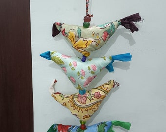 May include: A string of four colorful fabric birds with floral patterns, hanging from a brown wooden surface with a bell at the bottom.