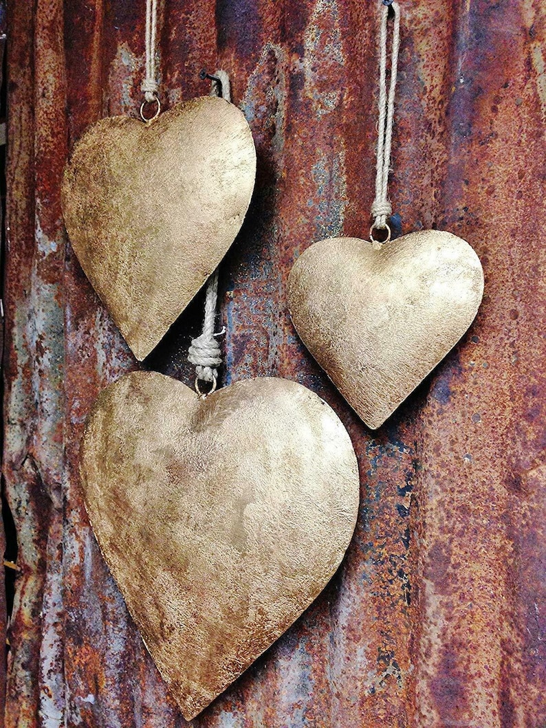 Op de afbeelding: Drie gouden metalen hartvormige ornamenten die aan touw hangen op een roestige metalen achtergrond.