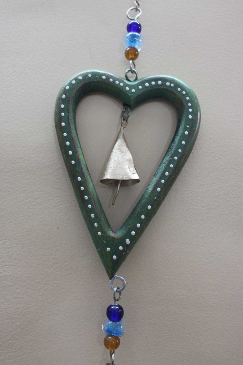Puede incluir: Un carill&oacute;n de madera verde en forma de coraz&oacute;n con lunares blancos y una campana dorada en su interior. El carill&oacute;n est&aacute; encordado con cuentas de vidrio azules y marrones.