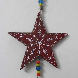 Puede incluir: Una estrella de madera roja con un dise&ntilde;o de copo de nieve blanco cuelga de una cuerda con cuentas de colores y una campana plateada.