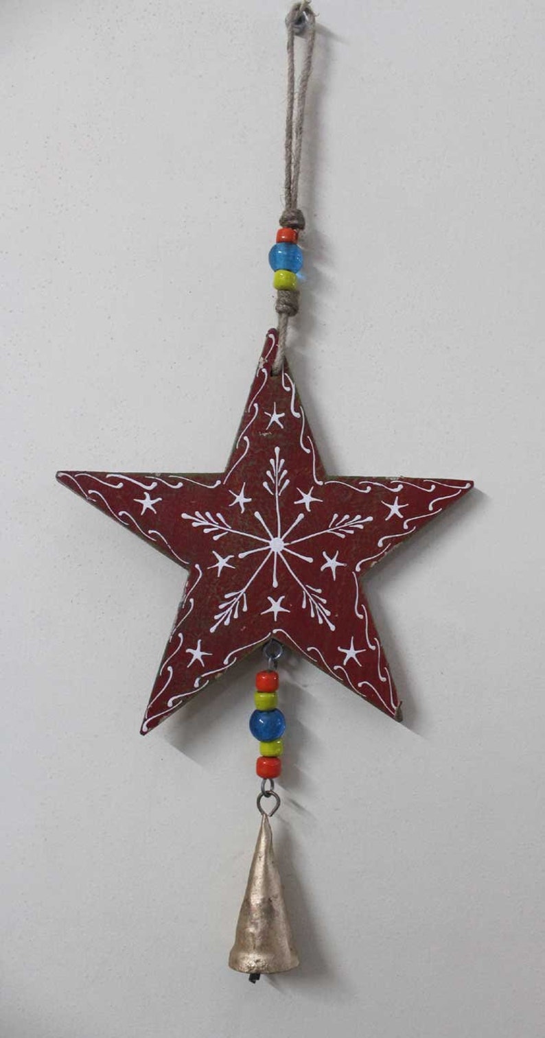 Puede incluir: Una estrella de madera roja con un dise&ntilde;o de copo de nieve blanco cuelga de una cuerda con cuentas de colores y una campana plateada.