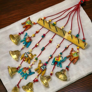 May include: A decorative wind chime featuring a bamboo bar, red cords, and colorful wooden beads. The chime includes small, painted elephant figurines in blue, yellow, and orange, along with brass bells. The design is set against a white background.