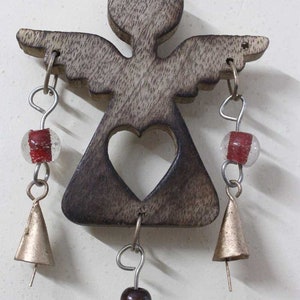 Puede incluir: Carill&oacute;n de viento de madera con forma de &aacute;ngel con un coraz&oacute;n recortado, cuentas marrones y blancas y campanas doradas.
