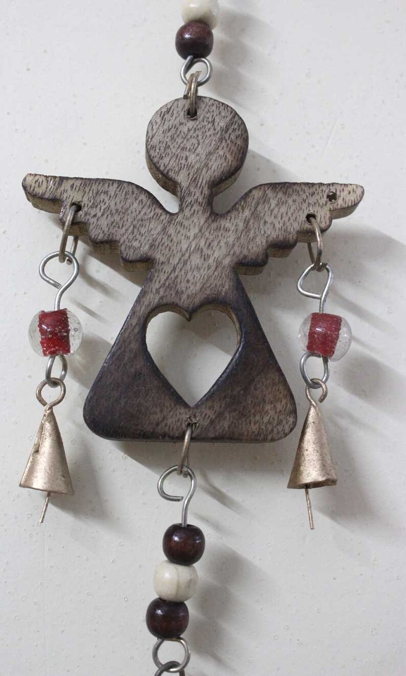 Puede incluir: Carill&oacute;n de viento de madera con forma de &aacute;ngel con un coraz&oacute;n recortado, cuentas marrones y blancas y campanas doradas.