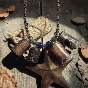 May include: Rustic necklace featuring a large, bronze-colored star pendant and several small, aged metal bells. The necklace has a black cord and is adorned with blue and green beads. A bundle of twine sits nearby on a weathered wooden surface.