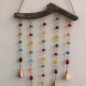 Op de afbeelding: Een houten windgong met vijf strengen kralen in rood, geel, groen en blauw. Elke streng eindigt met een kleine, gouden bel.