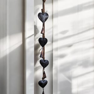 May include: A string of five black heart-shaped bells hanging from a brown rope. The bells are attached to the rope with small metal rings. The bells are hanging against a white wall.