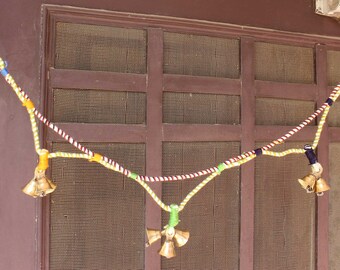 May include: A string of bells with colorful yarn, hanging from a brown wooden door with a wire mesh screen. The bells are gold and have a traditional design.