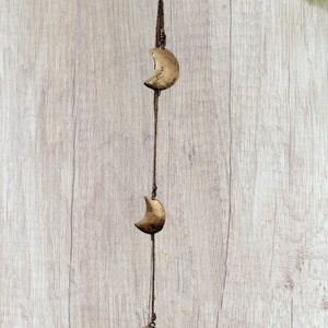 Puede incluir: Una cuerda marrón con tres colgantes de media luna de color dorado que cuelgan de ella. La cuerda está colgada sobre un fondo de madera clara.