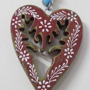 Puede incluir: Un adorno de madera en forma de coraz&oacute;n rojo con un dise&ntilde;o floral verde y blanco. El adorno est&aacute; colgado de una cadena plateada.