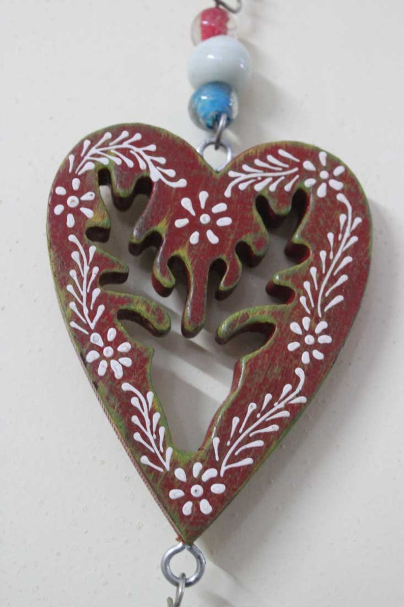 Puede incluir: Un adorno de madera en forma de coraz&oacute;n rojo con un dise&ntilde;o floral verde y blanco. El adorno est&aacute; colgado de una cadena plateada.