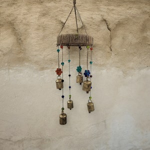 Puede incluir: Un carillón de madera con múltiples campanas colgando de él. Las campanas son de color dorado y tienen una variedad de formas. El carillón está decorado con cuentas de colores.