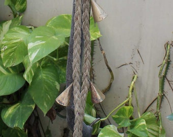 May include: A braided rope with a series of small, silver-colored bells hanging from it. The rope is hanging from a tree branch and is surrounded by green leaves.