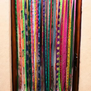 Può includere: Una colorata tenda per porta composta da strisce di tessuto verticali in vari modelli e colori, tra cui rosa, verde, blu e giallo. La tenda è appesa a un'intestazione in tessuto blu e presenta piccole nappe color oro nella parte inferiore di ogni striscia. Il telaio della porta è in legno.