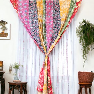 Puede incluir: Una cortina de patchwork colorida hecha de diferentes telas con patrones florales en tonos de rosa, amarillo, verde y azul. La cortina está atada hacia atrás en el centro y cuelga de una varilla blanca.