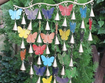 May include: A colourful butterfly wind chime with multiple tiers of bells and butterflies. The chime is made of metal and has a silver finish. The butterflies are painted in various colours, including red, blue, green, yellow, and orange. The chime is hanging from a chain and is suspended in front of a green bush.