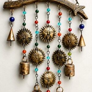 Puede incluir: Un colgante de pared con una rama de madera curvada que sostiene cinco caras de sol de metal y cinco campanas de latón. El conjunto está unido por cadenas adornadas con cuentas de vidrio multicolor en tonos azules, verdes, rojos y naranjas.