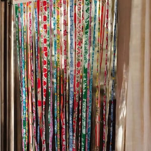 Pode incluir: Uma cortina decorativa composta por tiras de tecido verticais com vários padrões e cores. As tiras, adornadas com desenhos florais e geométricos em vermelho, azul, verde e amarelo, pendem de um cabeçalho branco com um padrão circular vermelho, criando um tratamento de janela vibrante.