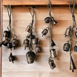May include: A collection of gold-toned metal bells hanging from twine on a wooden wall. The bells are of various sizes and shapes, with some having a more ornate design than others.