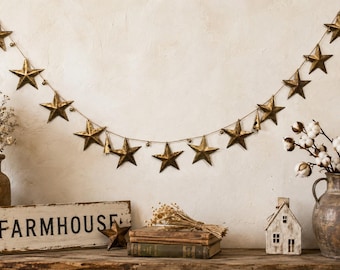 May include: A decorative garland featuring a series of metallic gold-toned stars strung along a thin wire, interspersed with small metal bells. The stars have a distressed finish and are arranged in a hanging arc against a neutral wall surface.