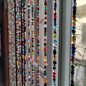 Op de afbeelding: Een kleurrijk kralen gordijn met strengen van verschillende kleuren en maten die aan een raam hangen. De kralen zijn gemaakt van glas, hout en andere materialen.