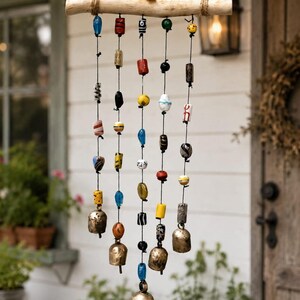 Op de afbeelding: Een hangend windgong met een horizontale, lichtgekleurde houten tak opgehangen aan een touw. Vijf verticale zwarte koorden hangen aan de tak, elk versierd met diverse kleurrijke glazen en keramische kralen en afgewerkt met een koperen bel.