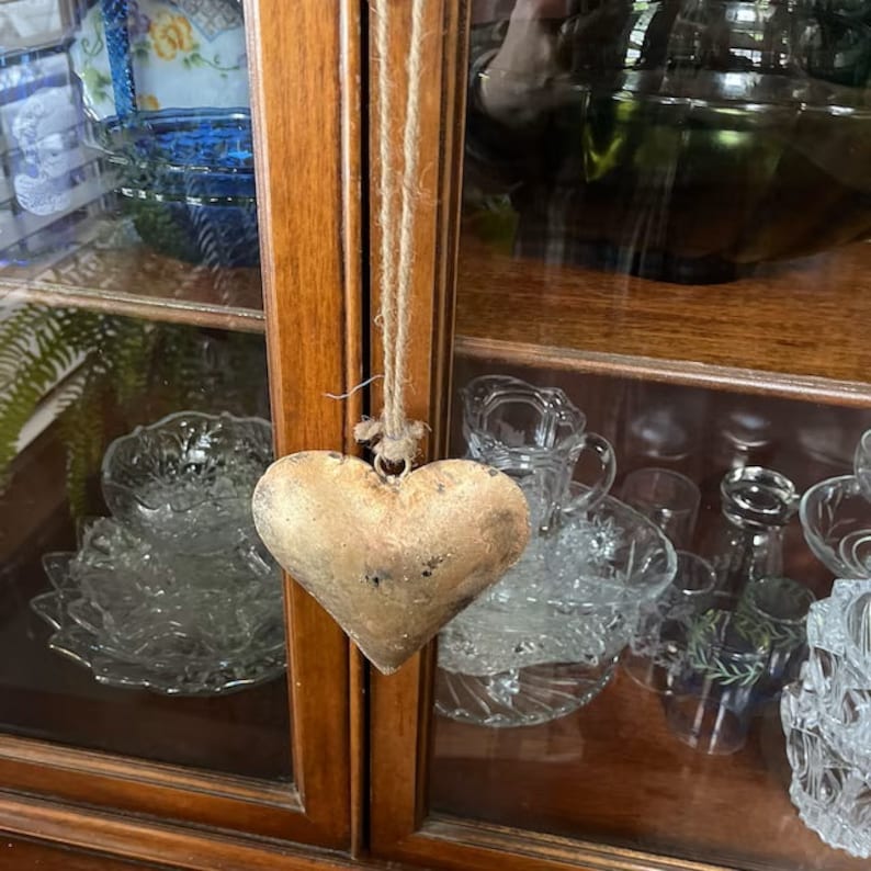 Op de afbeelding: Een houten kast met glazen deuren en planken. Een gouden hartvormig ornament hangt aan een touwtje in de kast. De planken zijn gevuld met glazen schalen en andere decoratieve items.