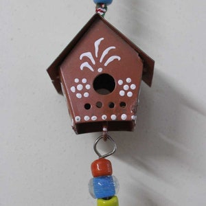 Puede incluir: Una casita de p&aacute;jaros de metal marr&oacute;n con detalles pintados en blanco cuelga de una cuerda de cuentas de colores. La casita de p&aacute;jaros tiene una peque&ntilde;a abertura redonda y un dise&ntilde;o decorativo en la parte delantera.